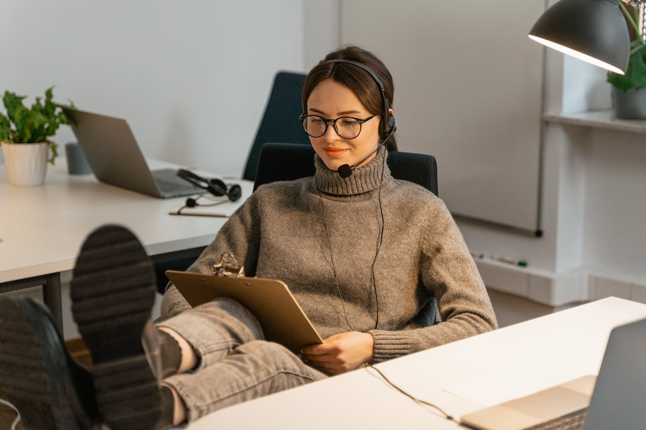 A smiling office worker with headphones, relaxed and reading a clipboard in a modern office.